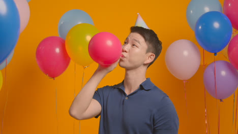 Studioporträt-Eines-Mannes-Mit-Partyhut,-Der-Seinen-Geburtstag-Feiert-Und-Einen-Ballon-In-Die-Luft-Sprengt-1
